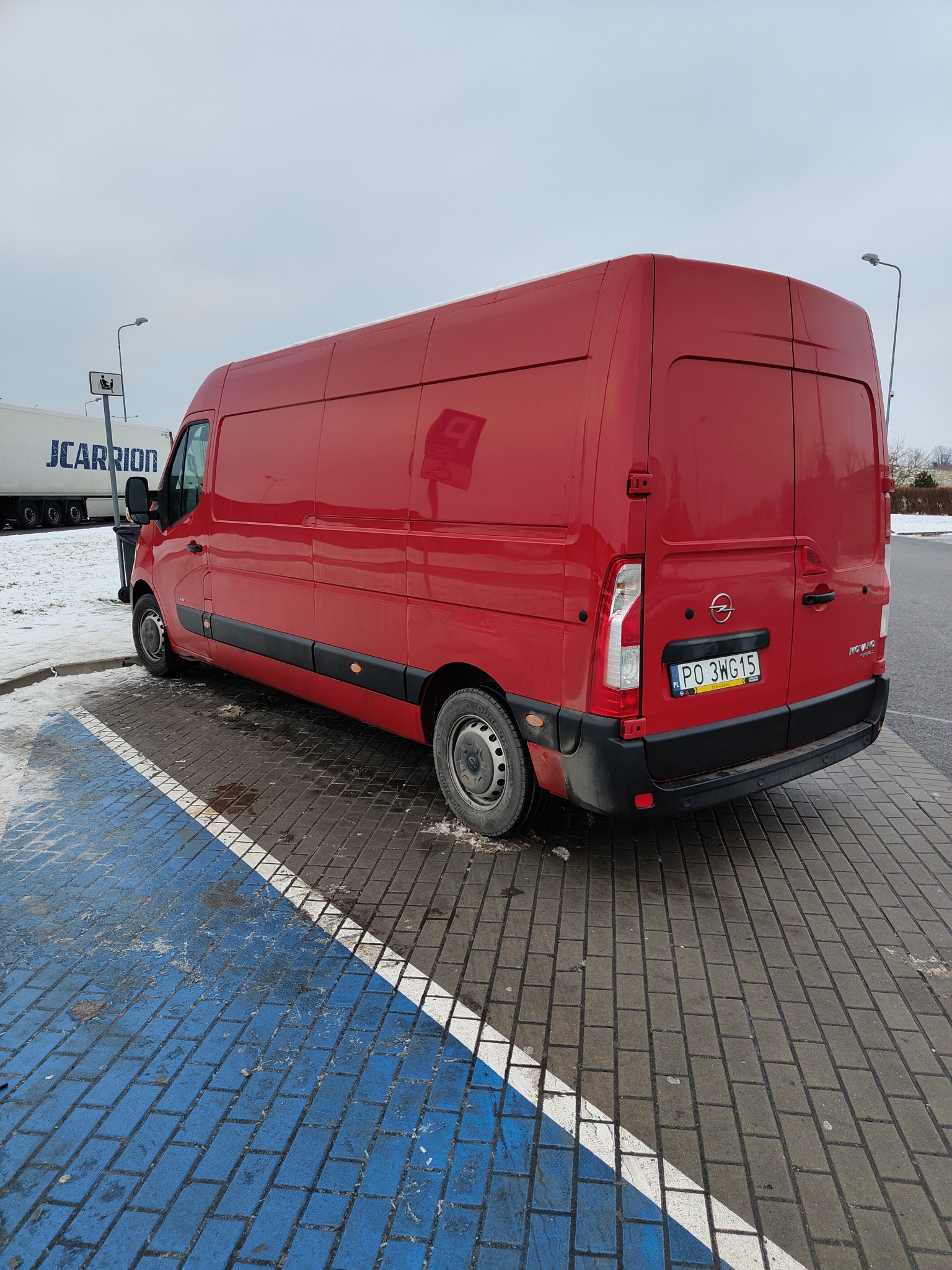 Czerwony Opel Movano zaparkowany na parkingu z niebieskimi liniami, widoczny z tyłu i boku, w tle biała naczepa J.Carrion.