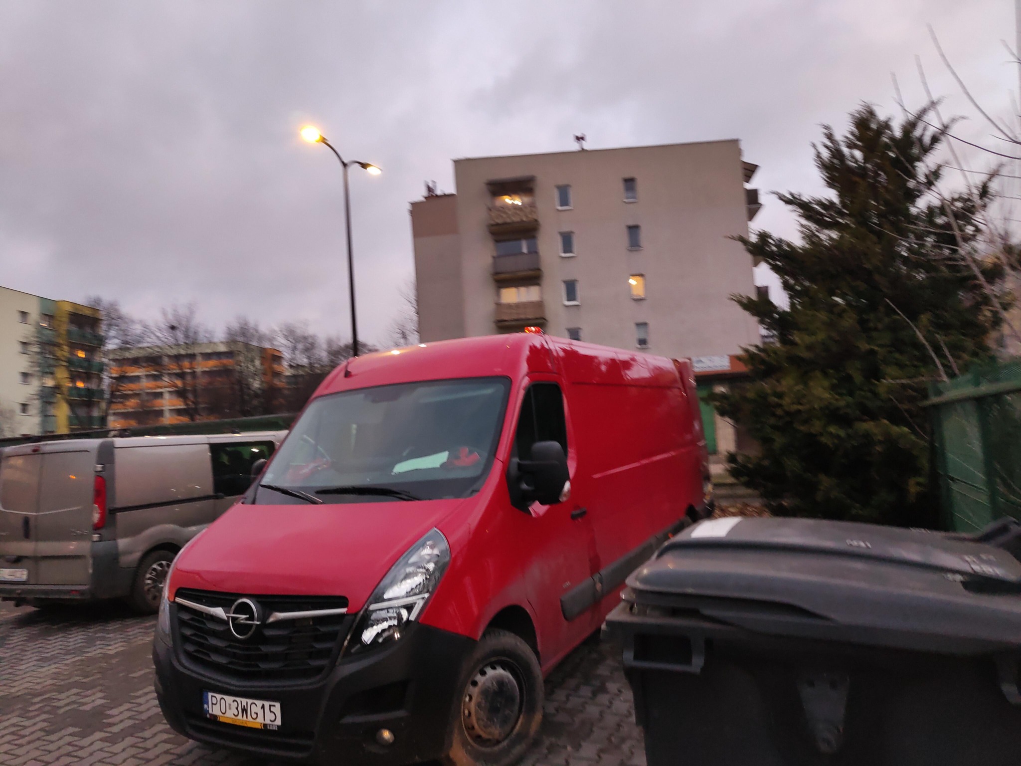 Czerwony bus Opel Movano na tle bloku mieszkalnego i pochmurnego nieba, zaparkowany obok pojemników na śmieci, widoczny numer rejestracyjny PO 3WG15.