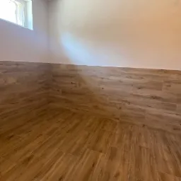 Wnętrze pomieszczenia z wyłożonymi panelami podłogowymi i częściowo ścianami, imitującymi drewno, małe okno w górnej części ściany.