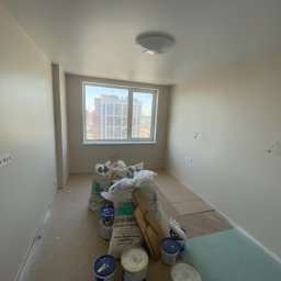 DACH INTERIOR STUDIO - Pokój w trakcie prac wykończeniowych, widoczne worki z zaprawą, puszki z farbą, fragment płyty kartonowo-gipsowej na podłodze, ściany pomalowane na jasny kolor, okno z widokiem na budynki.