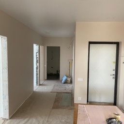 DACH INTERIOR STUDIO - Korytarz w trakcie remontu: zabezpieczona podłoga, ściany w neutralnym beżu, widoczne przygotowania do prac wykończeniowych przy ościeżnicach, narzędzia na piance izolacyjnej.