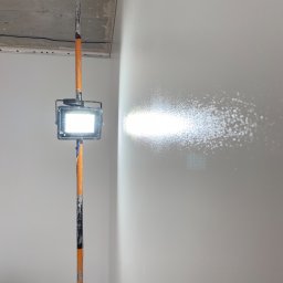 DACH INTERIOR STUDIO - Lampa robocza na teleskopowym stojaku oświetla świeżo pomalowaną, gładką ścianę w rogu pomieszczenia podczas prac wykończeniowych.