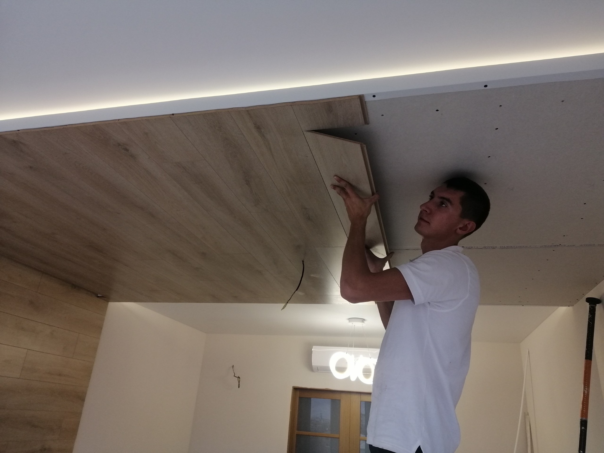 Mężczyzna montuje panele na suficie podwieszanym z płyty karton gips, widoczne oświetlenie LED podsufitowe i fragment ściany z panelami.