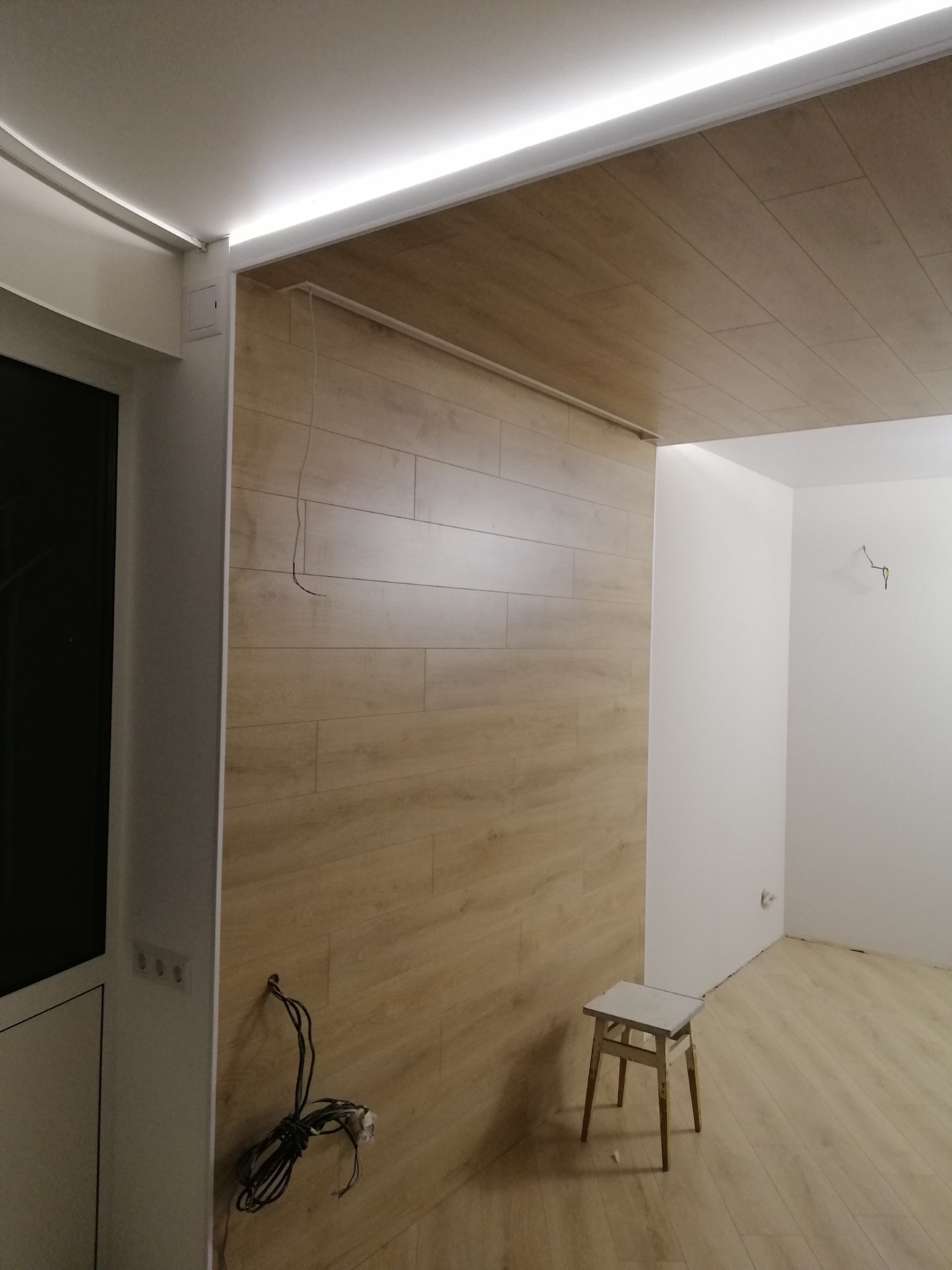 Wnętrze w trakcie wykańczania: ściana i sufit wyłożone panelami imitującymi drewno, widoczne wystające kable i mały stołek.