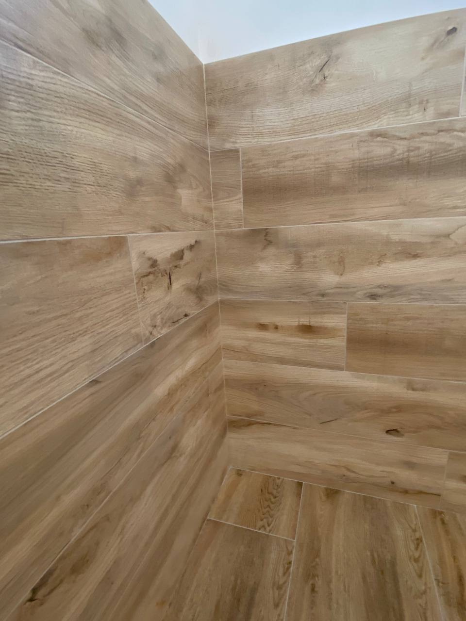 Wykończona ściana i podłoga w łazience wyłożona płytkami imitującymi drewno w odcieniach beżu i brązu, widok na narożnik.