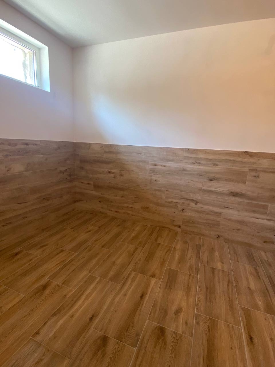 Wnętrze pomieszczenia z wyłożonymi panelami podłogowymi i częściowo ścianami, imitującymi drewno, małe okno w górnej części ściany.