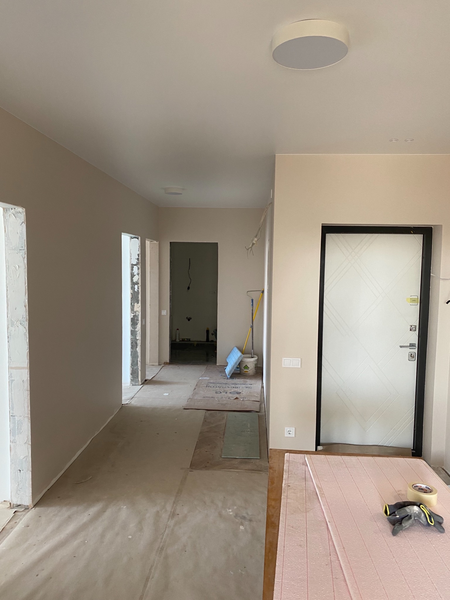 Korytarz w trakcie remontu: zabezpieczona podłoga, ściany w neutralnym beżu, widoczne przygotowania do prac wykończeniowych przy ościeżnicach, narzędzia na piance izolacyjnej.