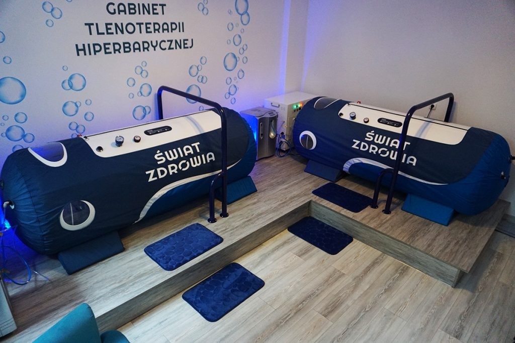 Dwie komory hiperbaryczne w gabinecie tlenoterapii, z napisem 'Świat Zdrowia', na podwyższeniu z drewnopodobnych paneli, z niebieskimi dywanikami i oświetleniem.