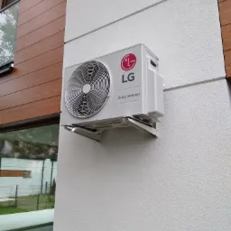 Zewnętrzna jednostka klimatyzacji LG Dual Inverter zamontowana na białej, teksturowanej ścianie budynku z elementami drewnianej okładziny.