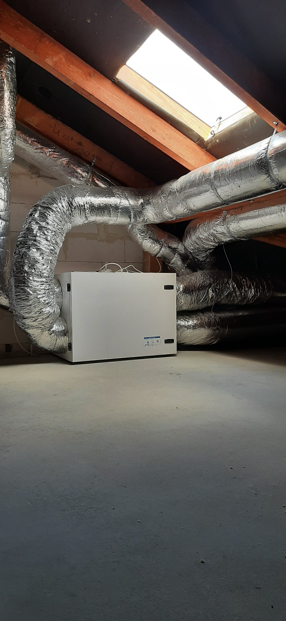 Biała centrala wentylacyjna z podłączonymi, izolowanymi przewodami wentylacyjnymi w pomieszczeniu na poddaszu z widocznym oknem dachowym.