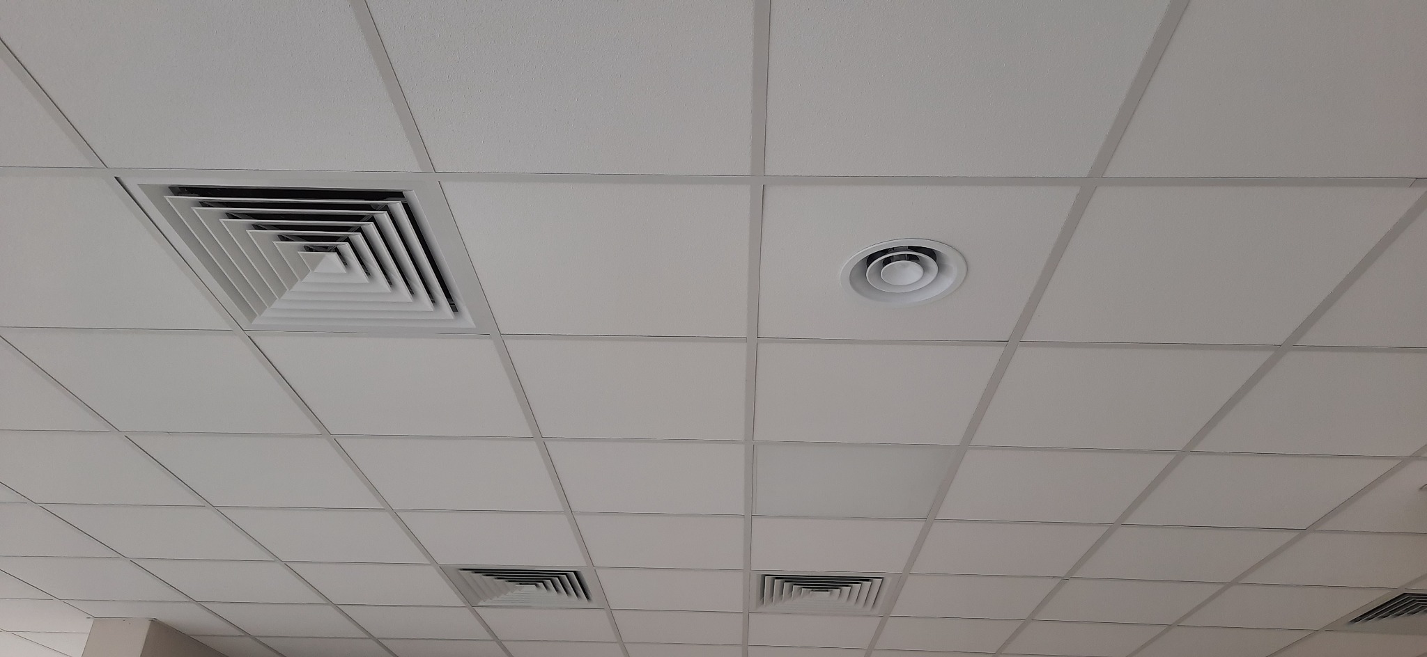 Sufit podwieszany z kwadratowymi kratkami wentylacyjnymi i okrągłym nawiewnikiem, widoczne z dołu pod kątem.