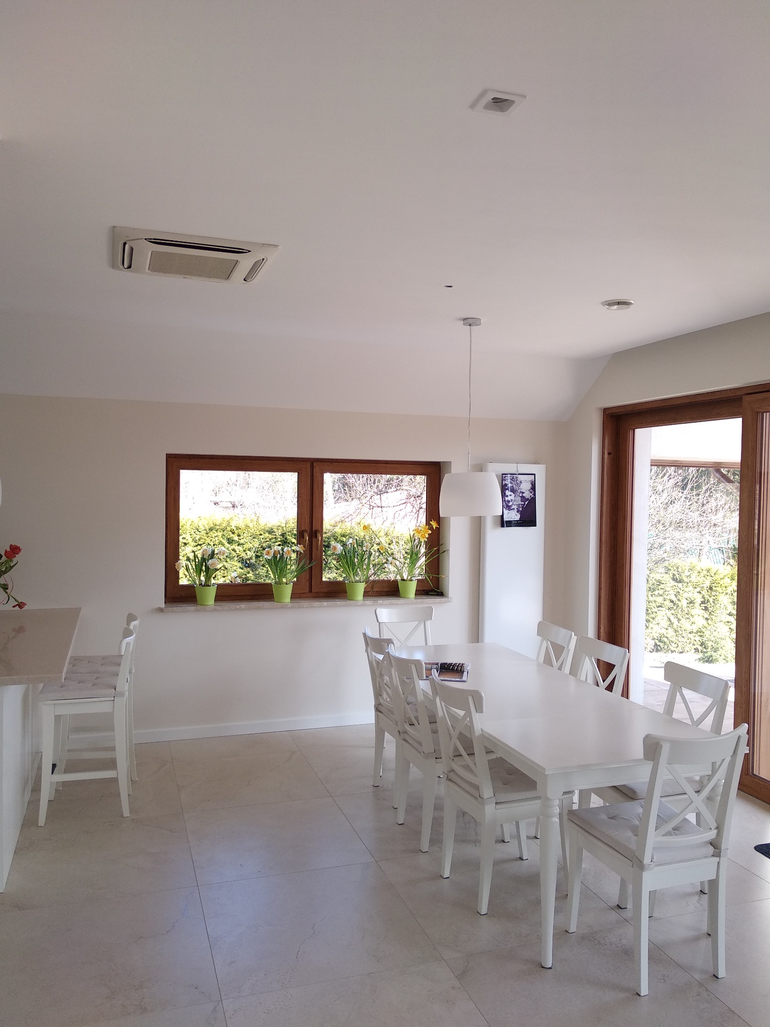 Wnętrze jasnej jadalni z białymi meblami i widoczną kasetą klimatyzacyjną w suficie. Na parapecie okiennym stoją doniczki z wiosennymi kwiatami.