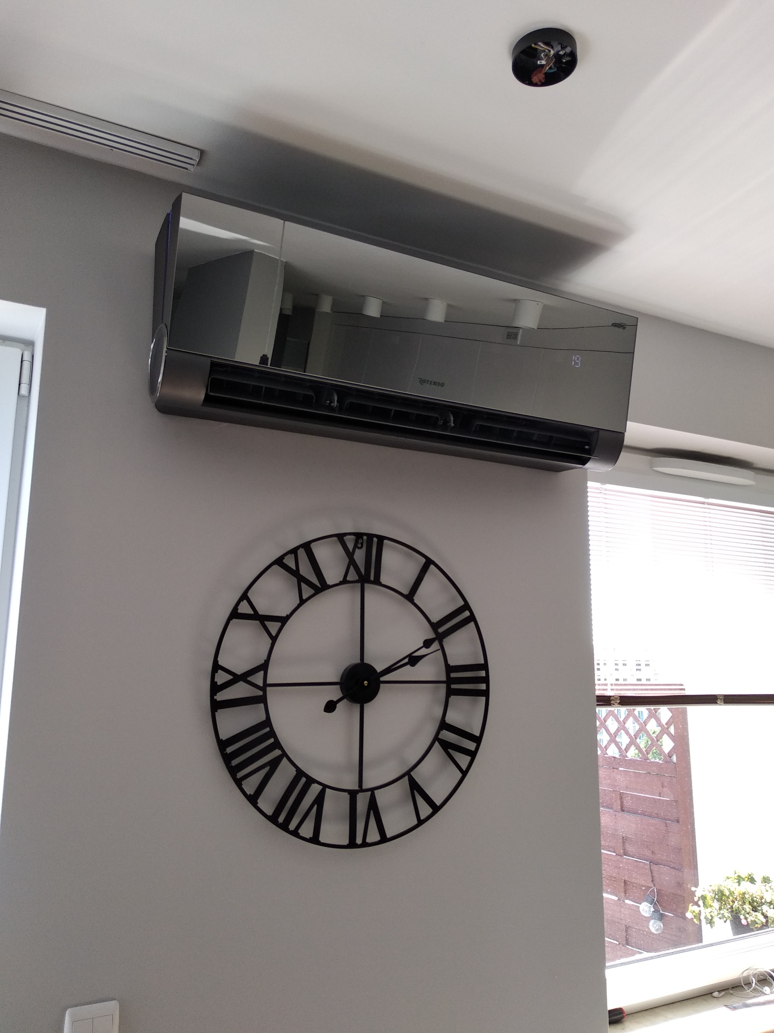 Szary klimatyzator ścienny w nowoczesnym wnętrzu, poniżej dekoracyjny zegar z rzymskimi cyframi, widok na okno z żaluzjami i roślinami.