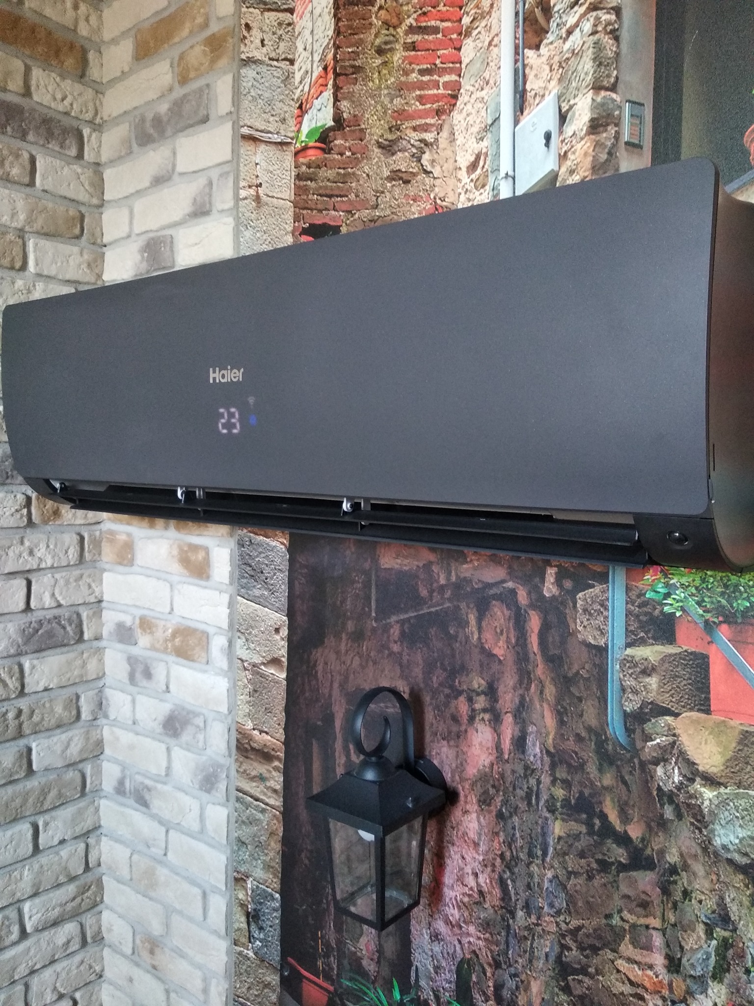 Czarny klimatyzator Haier zamontowany na ścianie z tapetą imitującą mur i latarnię, z wyświetlaczem wskazującym temperaturę 23 stopnie.