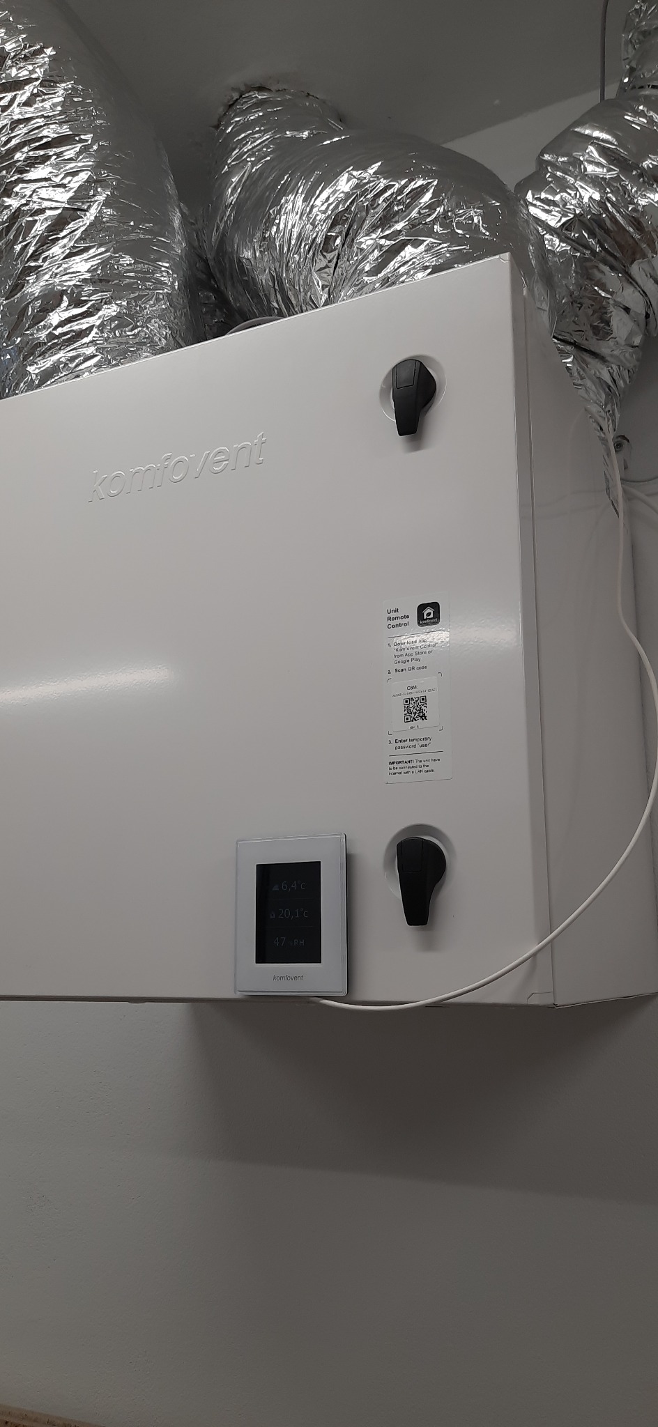 Centrala wentylacyjna Komfovent z widocznymi przewodami wentylacyjnymi w tle, wyświetlaczem temperatury i wilgotności oraz instrukcją obsługi na obudowie.