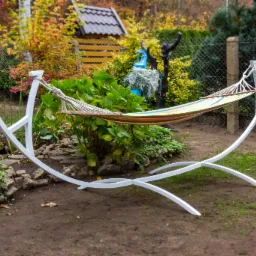 Biały, metalowy stelaż do hamaka z zawieszonym, kolorowym hamakiem w ogrodzie, na tle zieleni i jesiennych drzew.