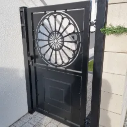 Czarna metalowa furtka z ozdobnym, okrągłym wzorem przypominającym rozetę, zamontowana przy wejściu na posesję. Widoczne detale mocowania i zamek.