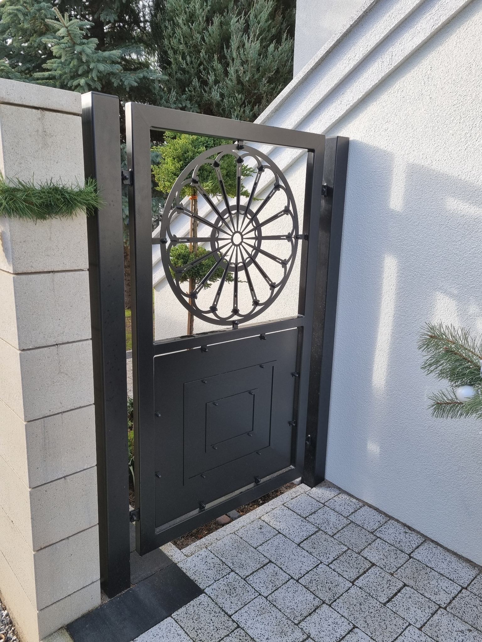 Metalowa furtka ogrodzeniowa w kolorze czarnym z ozdobnym, okrągłym wzorem w górnej części, zamontowana przy białym murze i brukowanym chodniku.