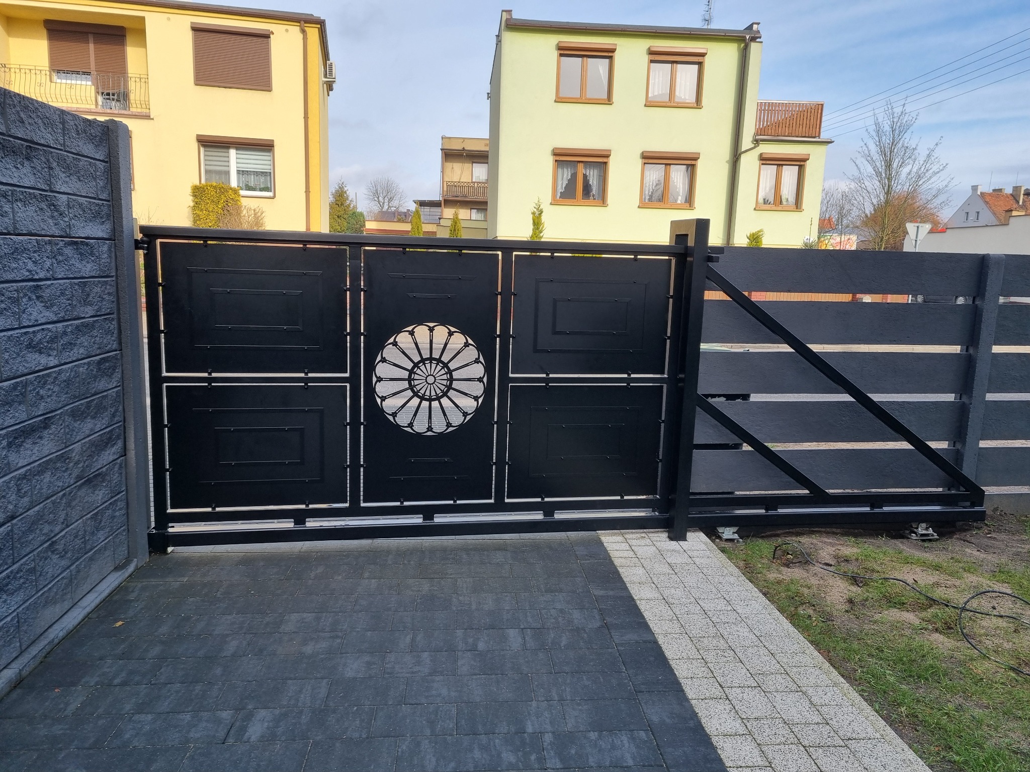 Czarna, przesuwna brama wjazdowa z metalu z dekoracyjnym, ażurowym motywem kwiatowym na tle domów jednorodzinnych i betonowego ogrodzenia.