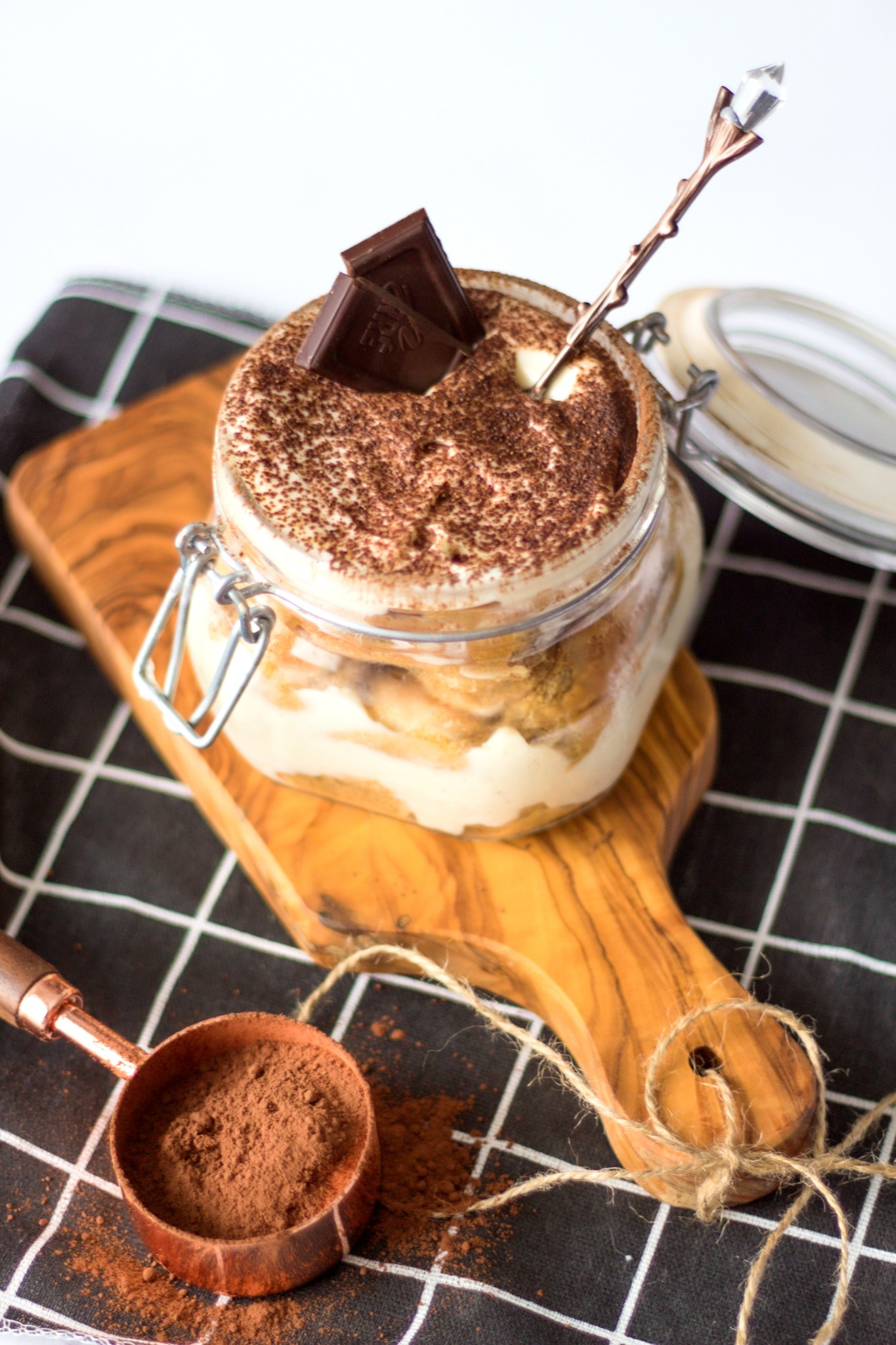 Tiramisu w słoiku z dwoma kostkami czekolady na wierzchu, ozdobna łyżeczka z kryształem, miarka kakao i drewniana deska na czarno-białej ściereczce.