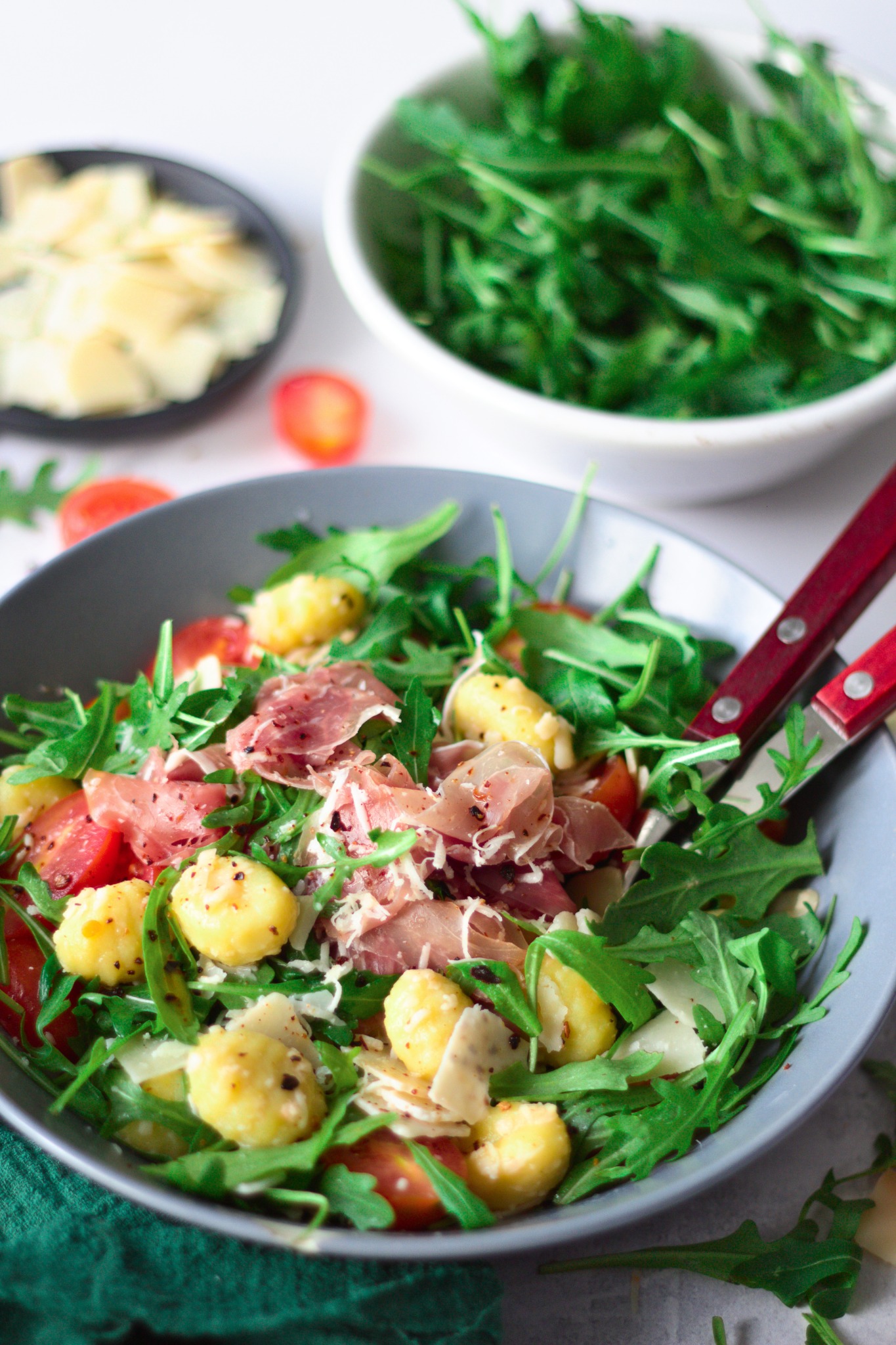 Sałatka z gnocchi, pomidorów, rukoli, szynki parmeńskiej i parmezanu w szarej misce, z dodatkową rukolą i serem w tle.