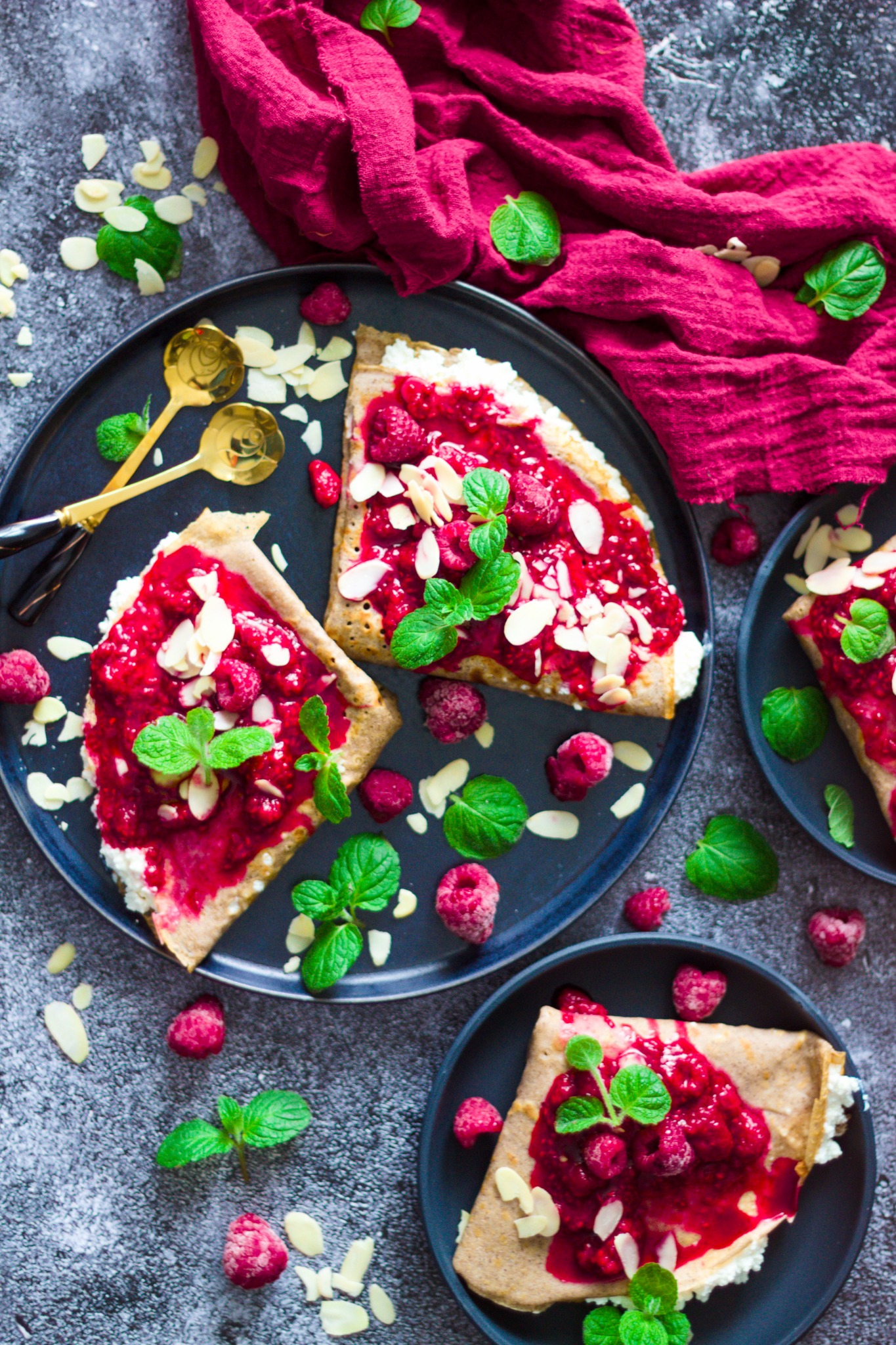 Naleśniki na ciemnych talerzach z twarożkiem, malinami, listkami mięty i płatkami migdałów, ułożone na szarym tle z dekoracyjną bordową tkaniną i złotymi łyżeczkami.