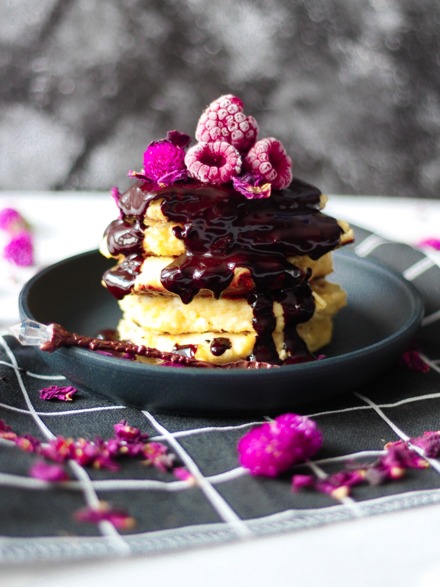 Stos naleśników polanych ciemną polewą, udekorowany malinami i jadalnymi kwiatami, podany na czarnym talerzu na tle szarej ściereczki w kratę.