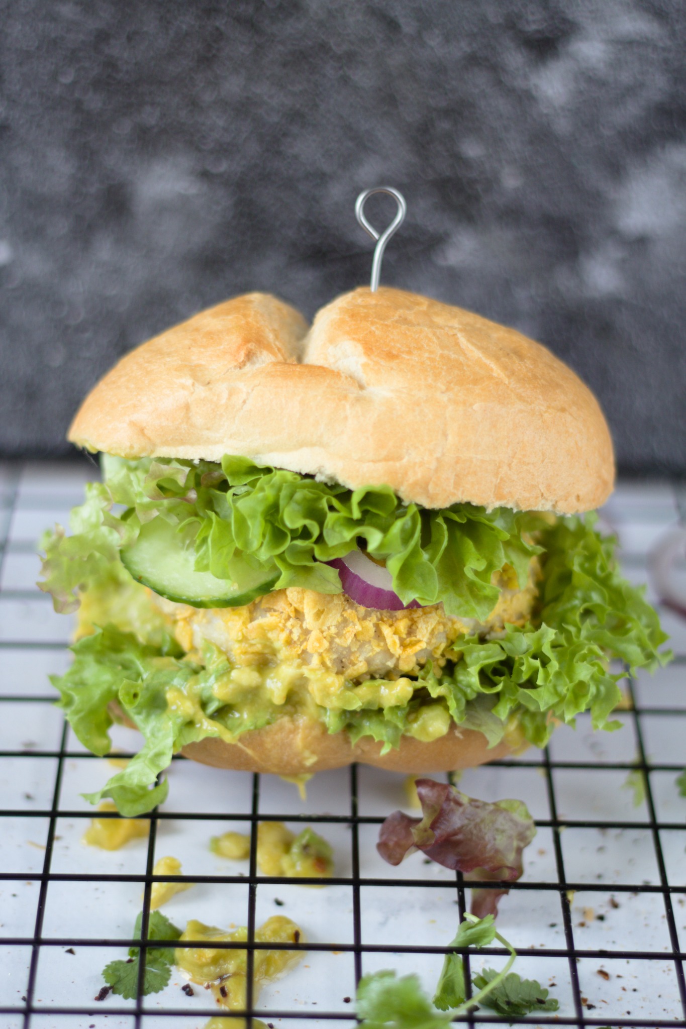 Kanapka z bułki pszennej z zieloną sałatą, plasterkiem ogórka, krążkiem czerwonej cebuli i kotletem z żółtą panierką, spięta metalowym szpikulcem, na białej kratce.