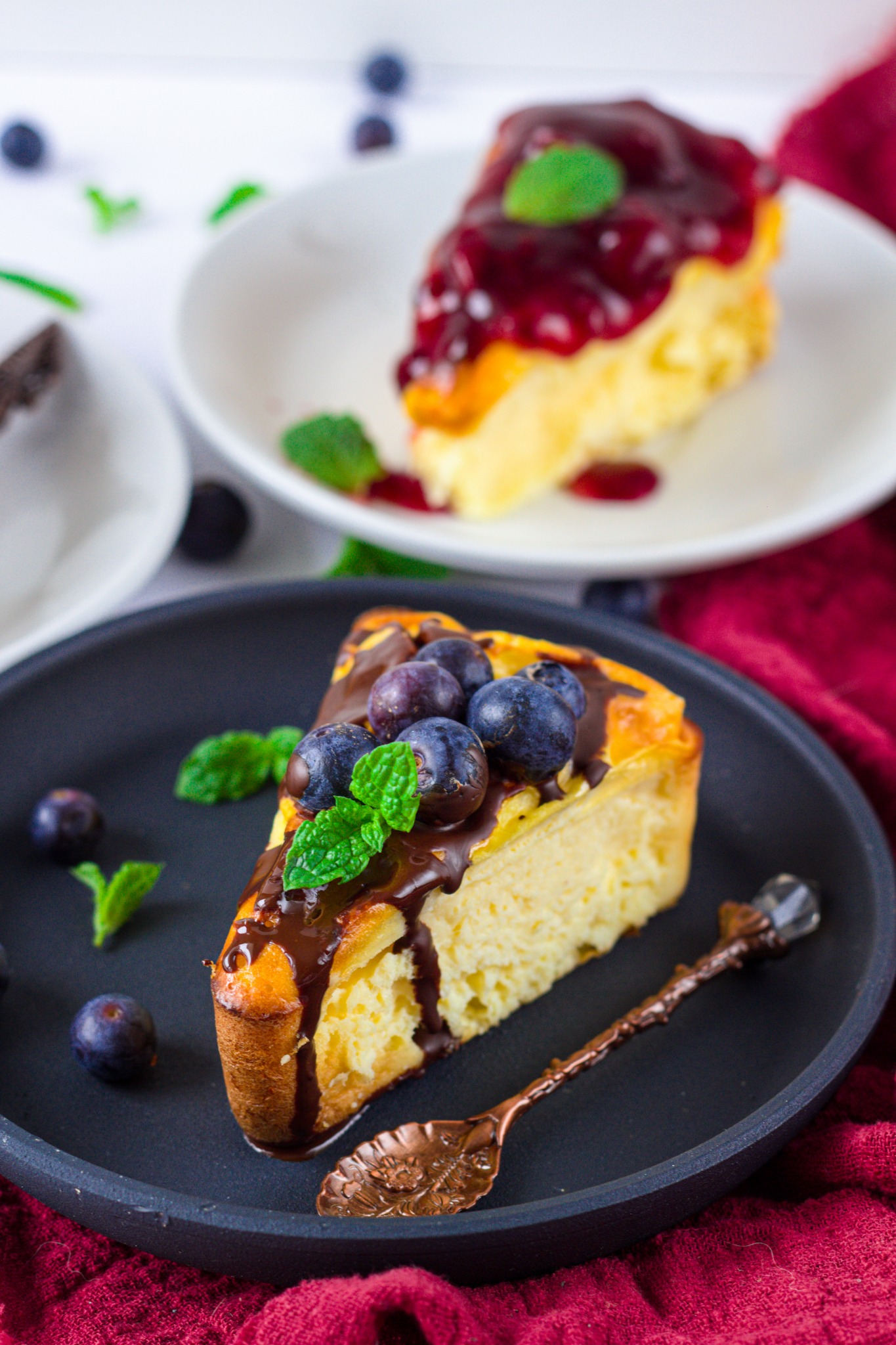 Kawałek sernika nowojorskiego z czekoladą, borówkami i miętą na czarnym talerzu obok dekoracyjnej łyżeczki, w tle drugi kawałek z sosem jagodowym.