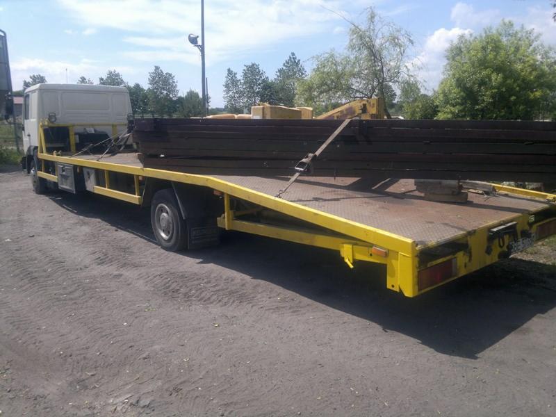 Żółta platforma niskopodwoziowa z ładunkiem belek stalowych zabezpieczonych pasem transportowym, widoczna na żwirowym placu w słoneczny dzień.