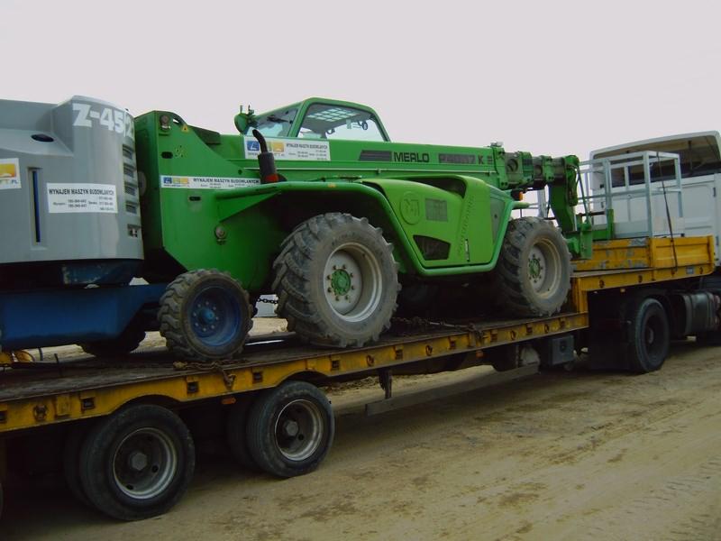 Zielona ładowarka teleskopowa Merlo P38.17 KE przewożona na żółtej niskopodwoziowej lawecie wraz ze sprzętem budowlanym w kolorze szarym.
