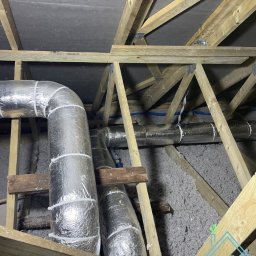 AIR4U - Izolowane przewody wentylacyjne w przestrzeni poddasza, widoczne drewniane elementy konstrukcyjne dachu oraz izolacja w postaci wełny mineralnej. W prawym dolnym rogu logo firmy.