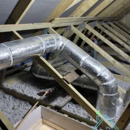 System kanałów wentylacyjnych w izolowanej przestrzeni poddasza, zabezpieczonych srebrną folią, widoczne elementy drewnianej konstrukcji dachu i włóknista izolacja.