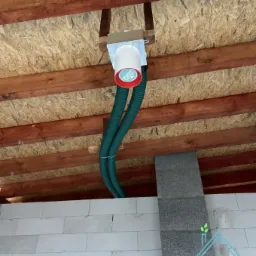 Centrala wentylacyjna z dwoma elastycznymi przewodami wentylacyjnymi, zamontowana pod drewnianym stropem na poddaszu budynku w trakcie budowy.