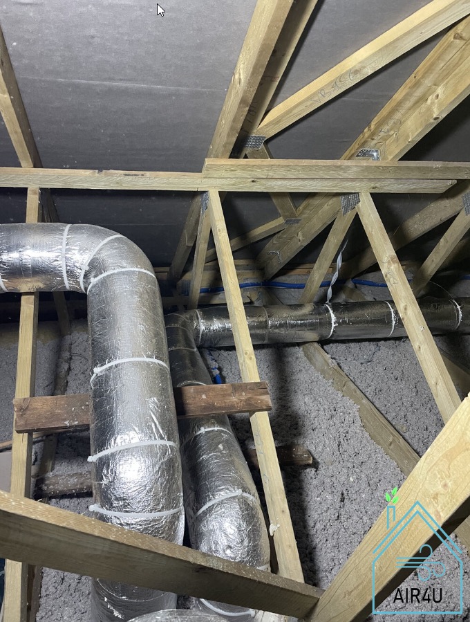 Izolowane przewody wentylacyjne w przestrzeni poddasza, widoczne drewniane elementy konstrukcyjne dachu oraz izolacja w postaci wełny mineralnej. W prawym dolnym rogu logo firmy.