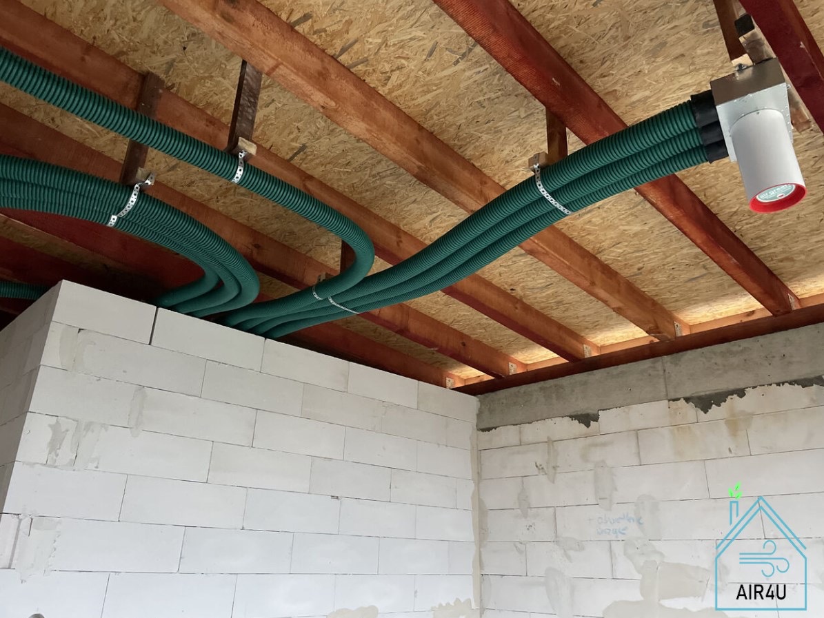 Zielone rury wentylacyjne przymocowane do drewnianego stropu w surowym wnętrzu z białymi ścianami z betonu komórkowego, widoczny fragment instalacji rekuperacji z anemostatem.