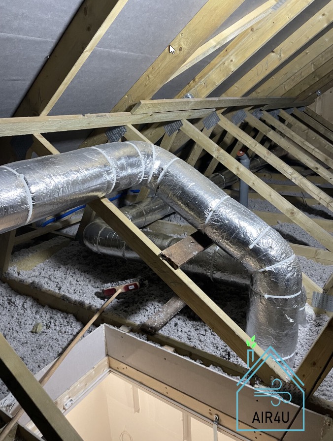System kanałów wentylacyjnych w izolowanej przestrzeni poddasza, zabezpieczonych srebrną folią, widoczne elementy drewnianej konstrukcji dachu i włóknista izolacja.