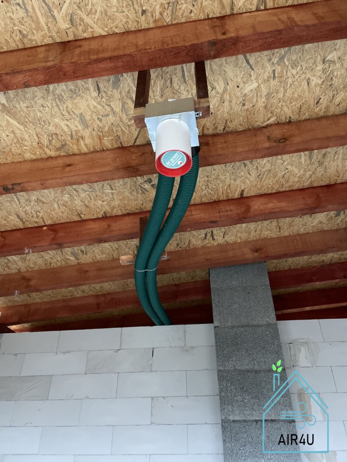 Centrala wentylacyjna z dwoma elastycznymi przewodami wentylacyjnymi, zamontowana pod drewnianym stropem na poddaszu budynku w trakcie budowy.