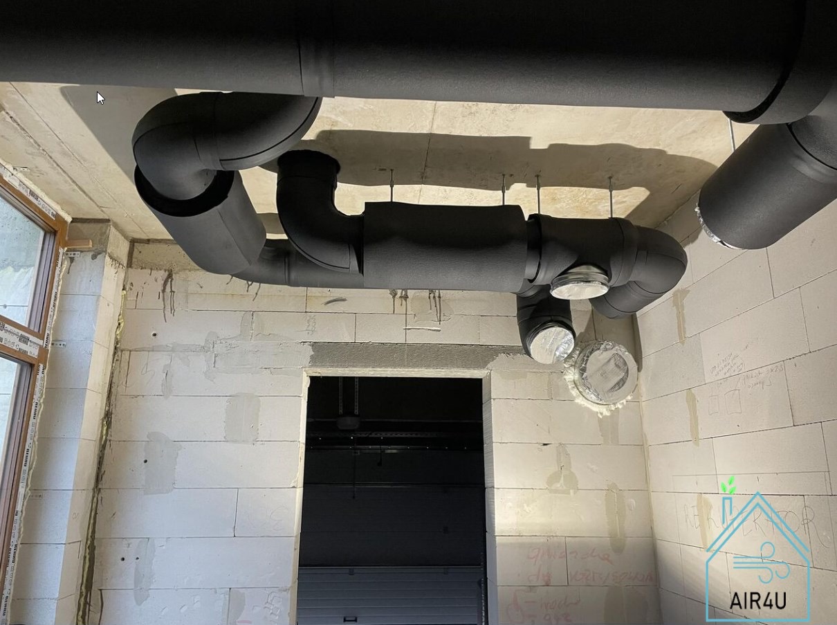 Czarne, zaizolowane rury wentylacyjne pod sufitem w surowym wnętrzu, widoczne elementy montażowe, ściany z białych pustaków, okno i otwór drzwiowy, logo firmy w prawym dolnym rogu.
