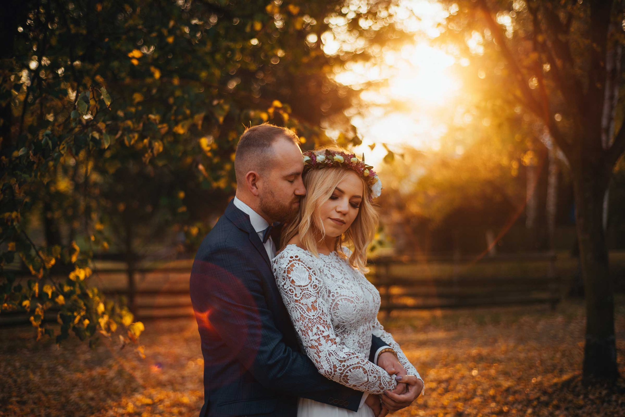 Para młoda w objęciach na tle jesiennego lasu oświetlonego zachodzącym słońcem, panna młoda w koronkowej sukni i wianku.