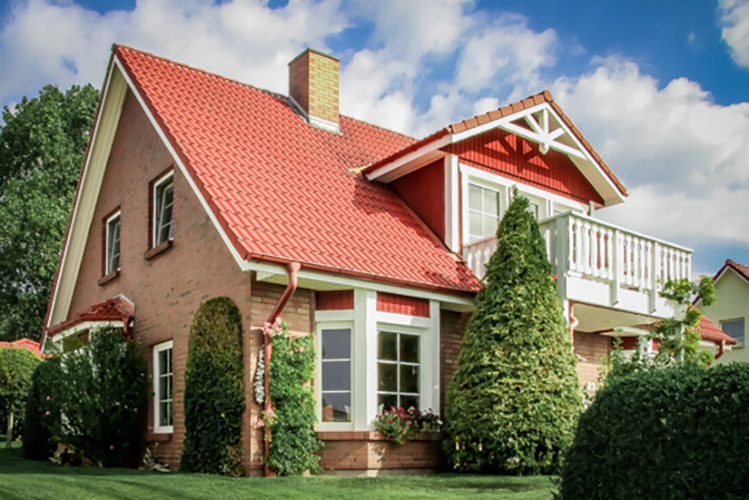 Dwupiętrowy dom z czerwonej cegły z czerwonym dachem i białym balkonem, otoczony zielonym trawnikiem i krzewami, pod błękitnym niebem z chmurami.