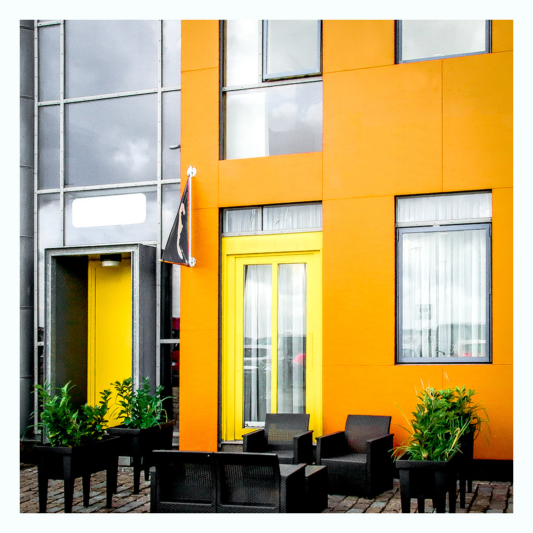Pomarańczowa fasada budynku z żółtymi drzwiami i oknami, czarną flagą, roślinami w donicach i czarnymi meblami ogrodowymi.