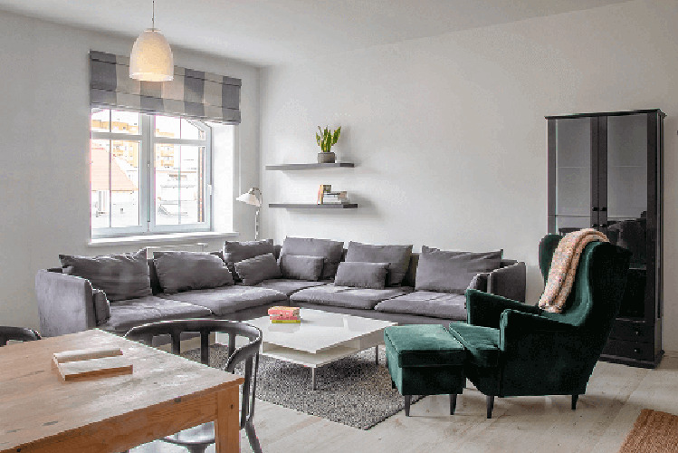 Jasne wnętrze salonu z szarą, narożną sofą, białym stolikiem kawowym, zielonym fotelem z podnóżkiem i drewnianym stołem jadalnym w rogu kadru. Na ścianie wiszą dwie półki z książkami i rośliną.