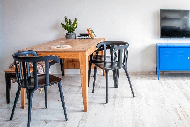 Jasne wnętrze z drewnianym stołem, czarnymi krzesłami, rośliną doniczkową, książkami i niebieską szafką pod telewizorem na białej podłodze.