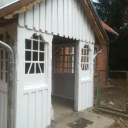 Biała, drewniana weranda z ozdobnymi okienkami, widoczna po remoncie, z elementami wykończeniowymi wciąż do poprawy, na tle ceglanego budynku.