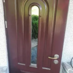 Ciemnobrązowe drzwi wejściowe z ozdobnym przeszkleniem, widok na zieleń ogrodu przez szybę, srebrna klamka i zamek, fragment ściany z szarego kamienia po prawej stronie.