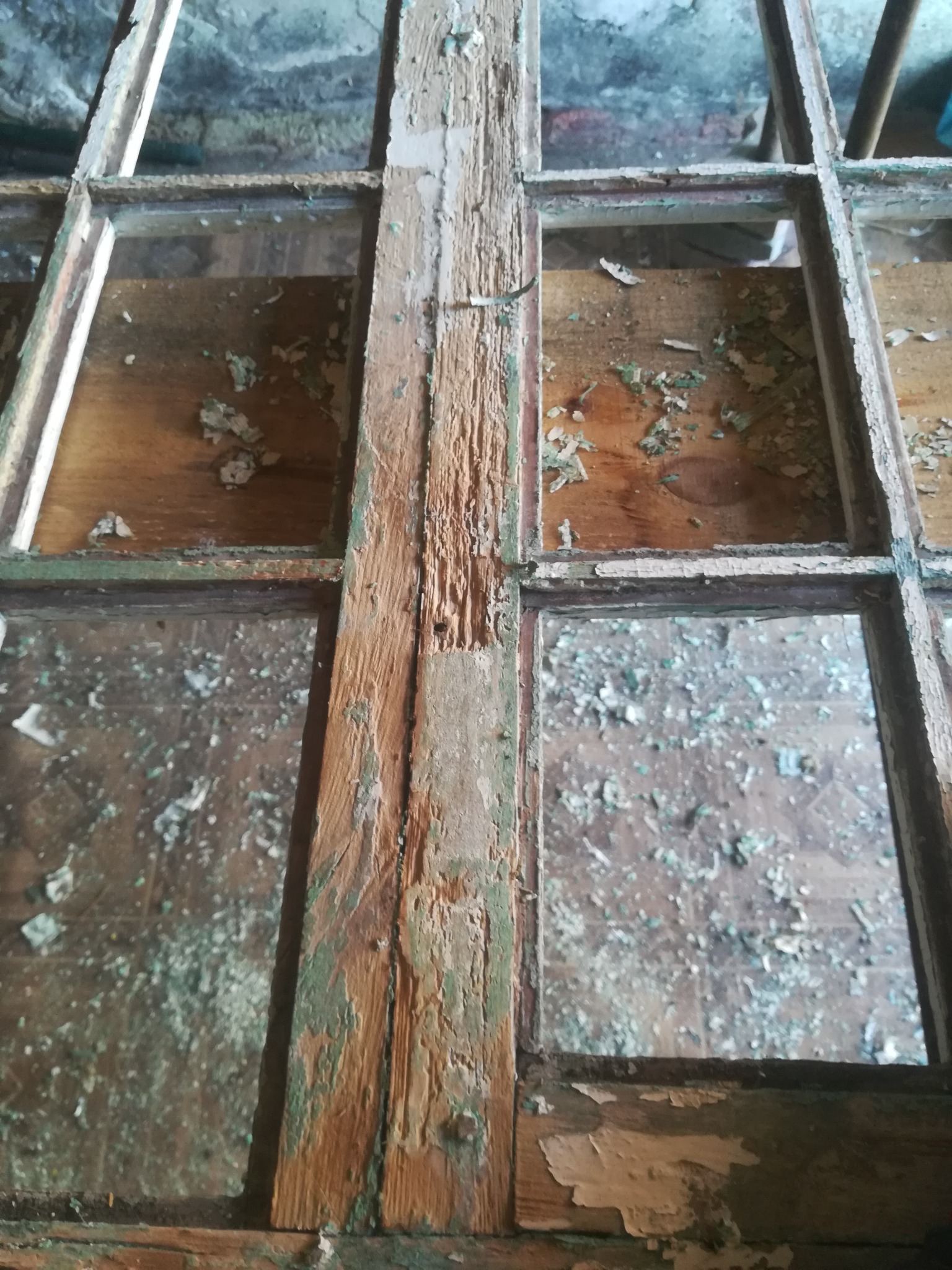 Stare, zniszczone okno drewniane z resztkami farby, widoczne ślady korozji i uszkodzenia drewna, szyby brudne, w tle widoczna ściana z cegły.