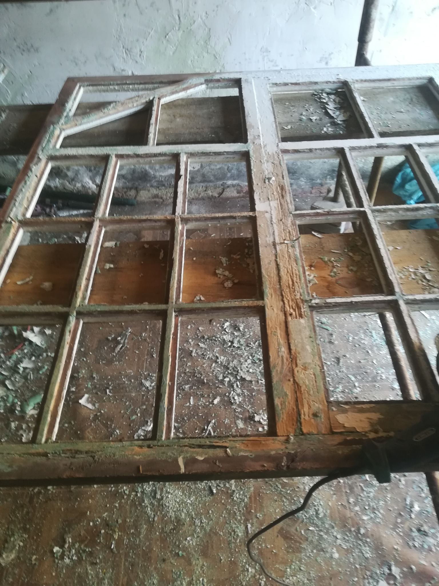 Stara, drewniana rama okienna z licznymi przegródkami, z resztkami farby i szkła, leżąca na podłodze pokrytej odpadami i resztkami farby, widoczne ślady użytkowania i zniszczenia.
