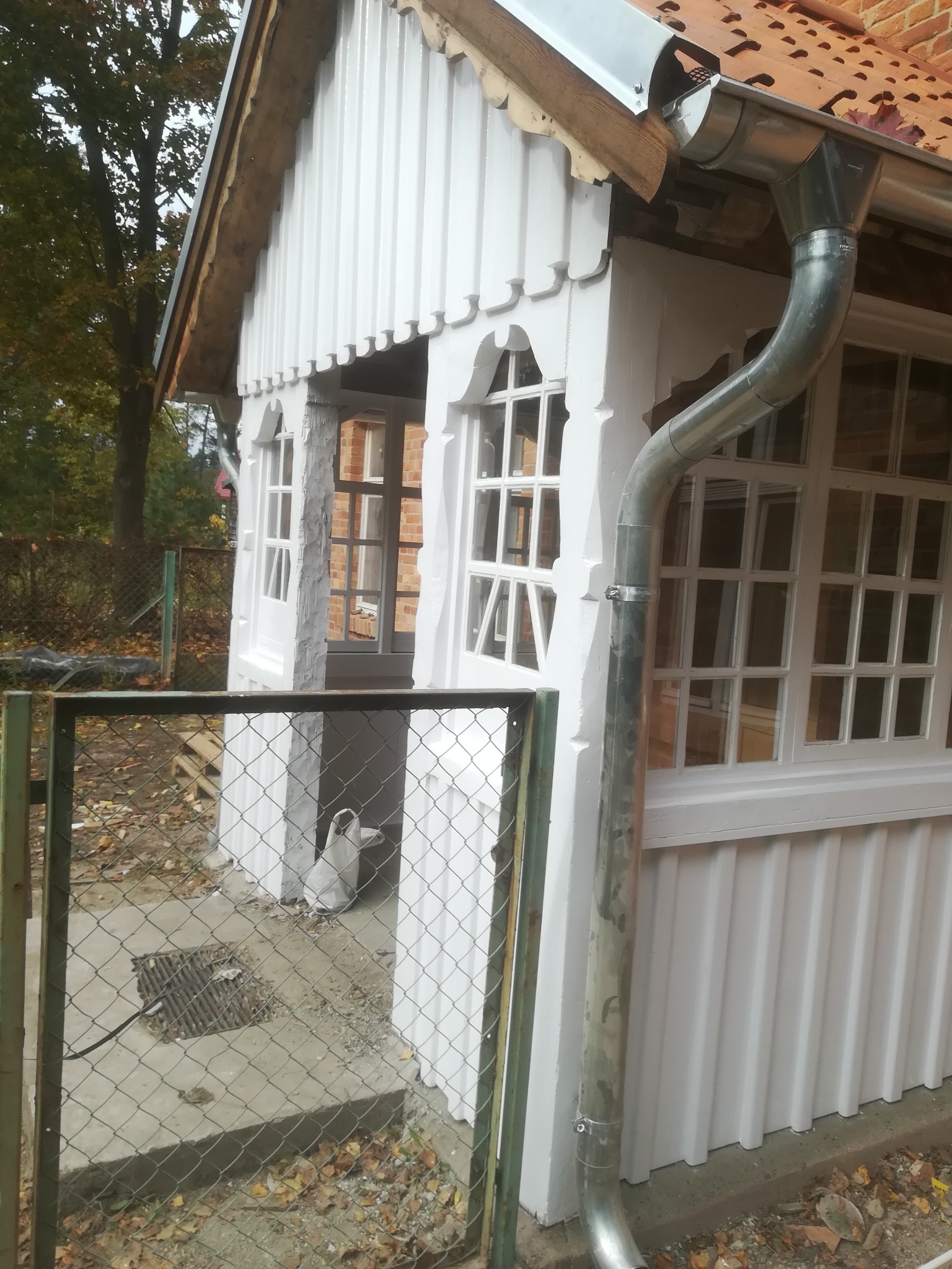 Biała, drewniana altana z dekoracyjnymi oknami i metalową rynną, widoczna przez ogrodzenie z siatki, z liśćmi na ziemi.