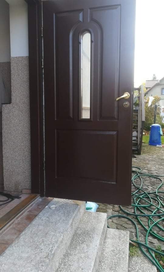 Ciemnobrązowe drzwi wejściowe z szybą, częściowo otwarte, widoczne betonowe schody wejściowe i zwinięty zielony wąż ogrodowy na brukowanym podłożu.